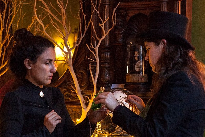 Against a backdrop of an ornately carved wooden bar decorated with barren branches, Josephine, dressed in dark lace with a locket at her neck, receives a drop of mysterious liquid from Doctor Vitae, who wears a top hat and gently holds a vial.