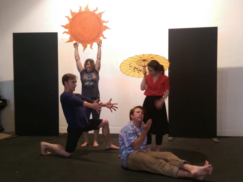 Four actors arranged in a beach scene: one holds a painted sun, one a yellow parasol, and one kneels with arms outstretched toward another who is sunning himself and relaxing.