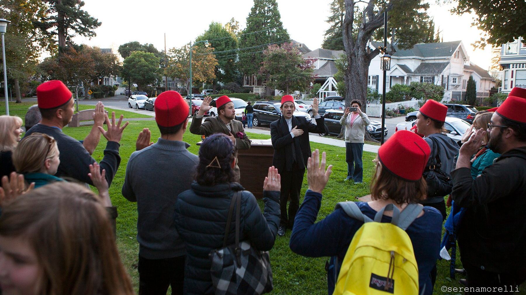 Audience and actors, many wearing red fezzes, recite a pledge holding one hand on their hearts and another in the air.